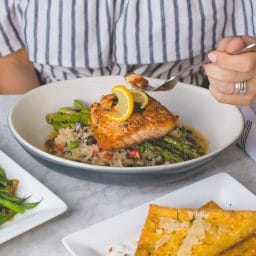 A person in front of a dinner plate of grilled fish and vegetables.