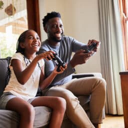 Little girl and her father laughing while playing video games together on a sofa in their living room at home